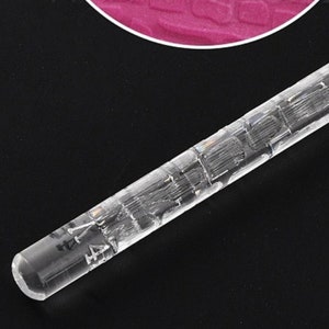 Puede incluir: Un rodillo cilíndrico transparente con marcas grabadas. El rodillo está hecho de un material transparente y tiene un extremo liso y redondeado. El diseño grabado añade un elemento decorativo a la herramienta de cocina.
