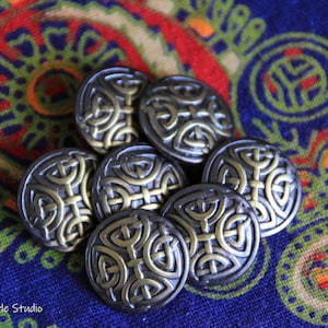 Könnte beinhalten: Sieben Metallknöpfe im antiken Stil mit einem keltischen Knotenmuster. Die Knöpfe sind silber- und goldfarben und liegen auf einem bunten Paisley-Stoffhintergrund. Jeder Knopf misst etwa 2,5 cm im Durchmesser.