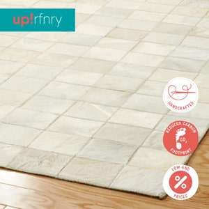 May include: Cream-colored, patchwork rug with a geometric pattern. The rug is made of rectangular pieces sewn together. The rug is on a wooden floor. The brand name "up! rfnry" is in the upper left corner. The rug is handcrafted with a reduced carbon footprint.