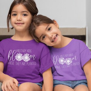 Op de afbeelding: Twee jonge meisjes dragen paarse T-shirts met de tekst "Military Kids Bloom Where They Are Planted" en witte paardenbloemontwerpen. De meisjes zitten op een trap en houden elkaars hand vast.