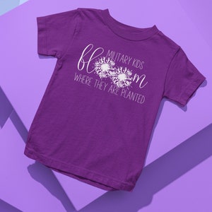 Op de afbeelding: Paars T-shirt met korte mouwen met witte tekst die "Military Kids Bloom Where They Are Planted" luidt. De tekst wordt omringd door drie witte paardenbloemontwerpen.
