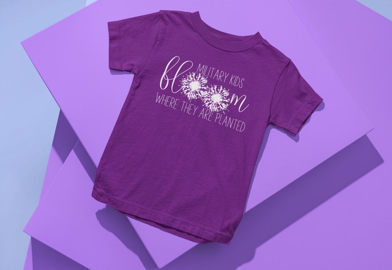 Op de afbeelding: Paars T-shirt met korte mouwen met witte tekst die "Military Kids Bloom Where They Are Planted" luidt. De tekst wordt omringd door drie witte paardenbloemontwerpen.