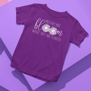 Op de afbeelding: Paars T-shirt met korte mouwen met witte tekst die "Military Kids Bloom Where They Are Planted" luidt. De tekst wordt omringd door twee witte paardenbloemontwerpen.