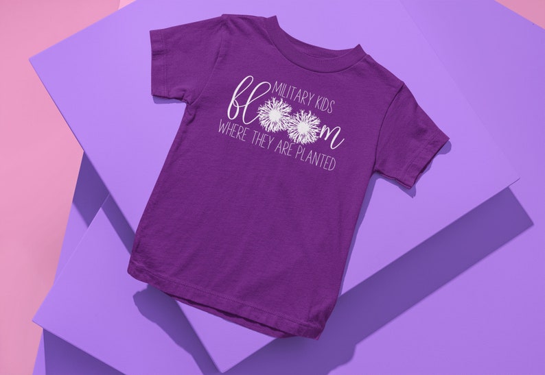 Op de afbeelding: Paars T-shirt met korte mouwen met witte tekst die "Military Kids Bloom Where They Are Planted" luidt. De tekst wordt omringd door twee witte paardenbloemontwerpen.