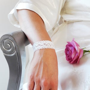 May include: A white lace bracelet worn on a person's wrist. The bracelet is delicate and features a simple design. The person is wearing a white robe and a pink rose can be seen in the background.