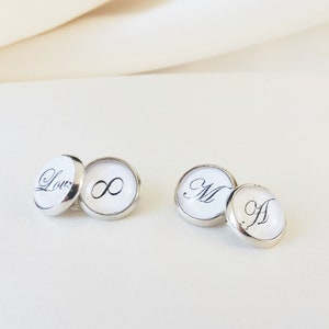 Può includere: Gemelli d'argento con facce bianche. Un paio presenta la parola "Love" e un simbolo di infinito in scrittura nera. L'altro paio mostra le lettere "M" e "A" in scrittura nera. I gemelli sono rotondi con una finitura lucida.