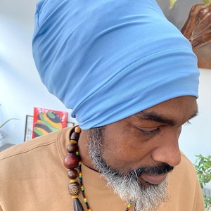 Puede incluir: Un gorro azul claro hecho de tela suave. El gorro lo lleva una persona con una camisa marrón y un collar de cuentas de madera. La persona tiene barba gris y está mirando hacia abajo.