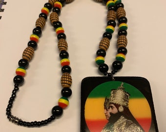 May include: A necklace with a large rectangular pendant. The necklace features black, red, yellow, and green beads, with wooden barrel-shaped beads. The pendant has a black border and a portrait with red, yellow, and green background.