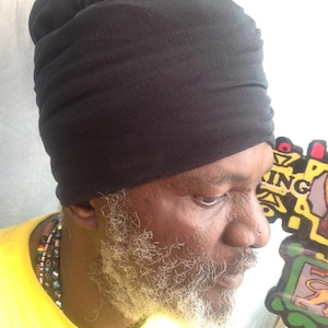 May include: A black turban is worn by a person with a yellow shirt and a beaded necklace. The person has a grey beard and is looking down. The background is a light colour.