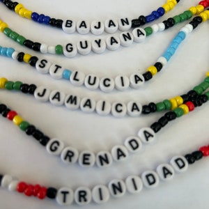 Może przedstawiać: Zbliżenie kilku naszyjników z koralików. Każdy naszyjnik zawiera nazwę innego kraju zapisaną białymi koralikami z literami. Koraliki są w różnych kolorach, w tym żółtym, niebieskim, zielonym i czerwonym. Nazwy obejmują: Bajan, Gujana, St. Lucia, Jamajka, Grenada i Trynidad.