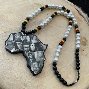 Può includere: Una collana con un ciondolo a forma di Africa, ornato con ritratti in bianco e nero di personaggi storici. La collana ha una fila di perle finte bianche, perline nere e accenti di perline rosse, gialle e verdi. È appoggiata su una superficie marrone chiaro.