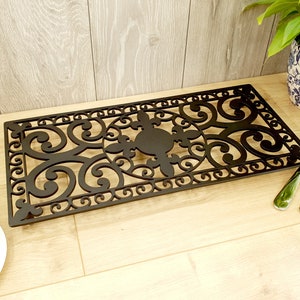 May include: Black metal doormat with a decorative, intricate design featuring swirls and flourishes. The doormat is rectangular and has a slightly raised edge.