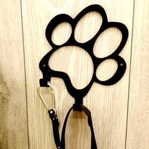 May include: Black metal paw print hook mounted on a wood wall with a black leash and silver carabiner hanging from it.