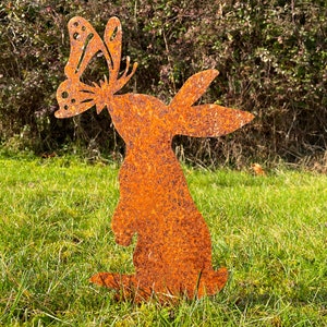 May include: A rusty metal garden sculpture of a rabbit with a butterfly on its head. The sculpture is standing on a bed of green grass.