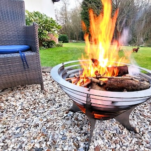 May include: A metal fire pit with a bright orange and yellow fire burning inside. The fire pit is surrounded by a gravel surface, with a wicker chair and a grassy backyard in the background.