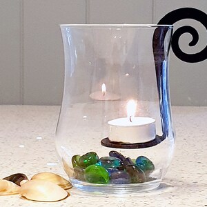 May include: A clear glass candle holder with a black metal swirl handle. The holder contains tealight candles, green and blue glass stones, and two white seashells.
