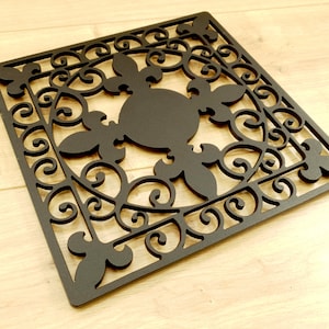 May include: A black metal decorative doormat with a square shape and an intricate floral design. The design features a central circle with four fleur-de-lis.