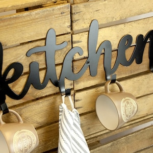 May include: Black metal kitchen sign with the word "kitchen" in a cursive font. The sign has three hooks, two of which are holding brown mugs with gold lettering. The third hook is holding a striped kitchen towel.