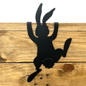 May include: A black metal bunny silhouette decoratively hanging on a wooden surface. The bunny is depicted mid-leap, with its ears and legs extended. The wooden surface has a natural, rustic appearance.