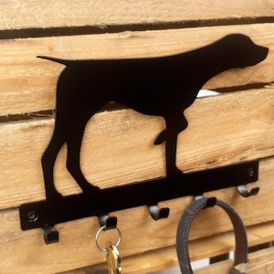 May include: Black metal key holder shaped like a dog silhouette with three hooks. The key holder is mounted on a wooden surface.