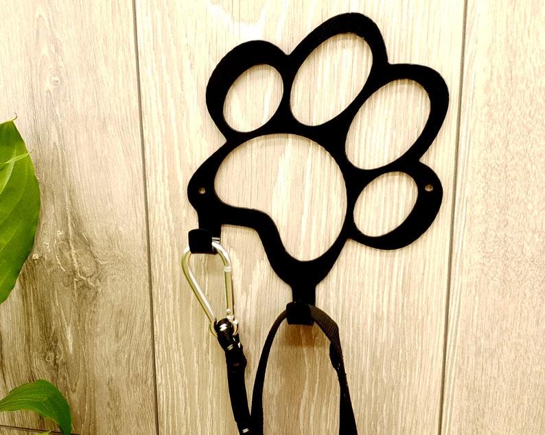 May include: Black metal paw print hook mounted on a wood wall with a black leash and silver carabiner hanging from it.