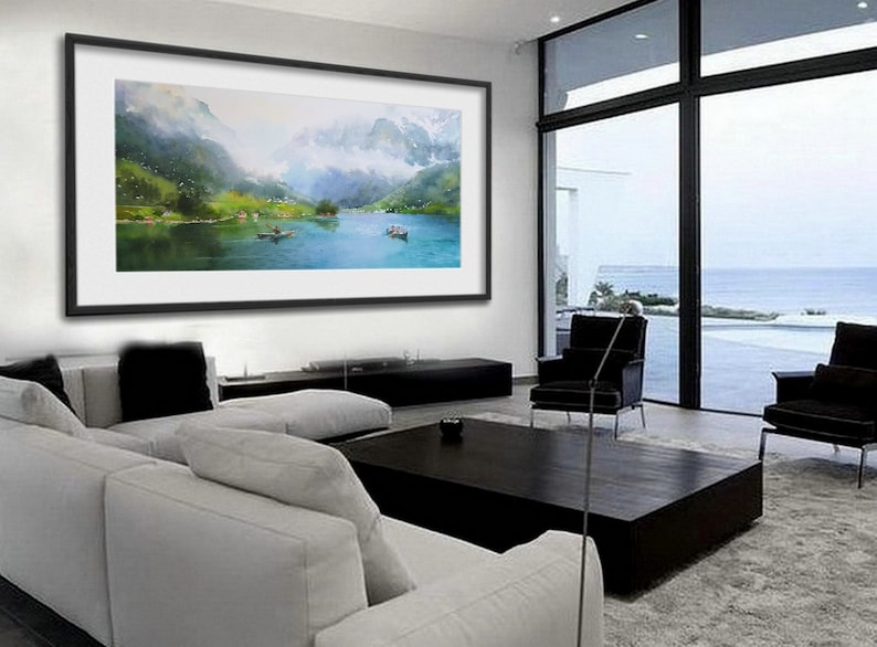 May include: A framed watercolor painting of a mountain lake scene with boats and a misty background. The painting is hanging on a white wall in a modern living room with a white couch, black coffee table, and large windows.