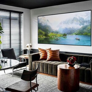 May include: A living room interior with a dark green sofa, a brown wooden coffee table, and a large framed watercolor painting of a mountain lake scene. The painting is hanging on a white wall above the sofa. The room has a large window with blinds and a black chair.