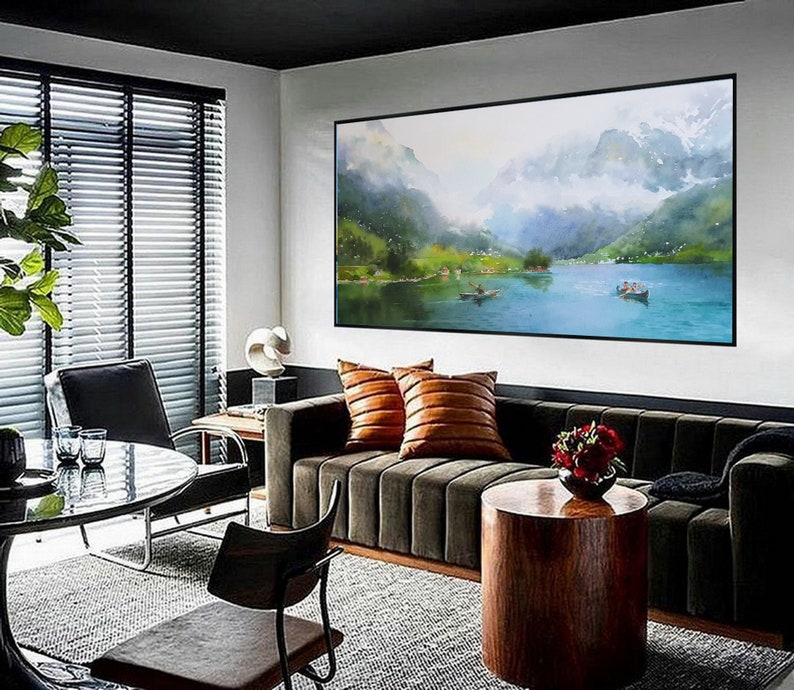 May include: A living room interior with a dark green sofa, a brown wooden coffee table, and a large framed watercolor painting of a mountain lake scene. The painting is hanging on a white wall above the sofa. The room has a large window with blinds and a black chair.