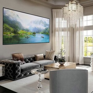 May include: A living room interior with a large framed watercolor painting of a lake and mountains, a gray tufted sofa with pillows, a gold coffee table, a clear glass side table, and a gray armchair.