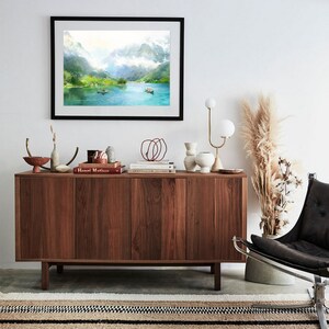 May include: A framed watercolor painting of a lake surrounded by mountains. The painting is hanging on a white wall above a wooden cabinet. The cabinet is decorated with books, vases, and other decorative items. A black leather chair with a metal frame sits in front of the cabinet.