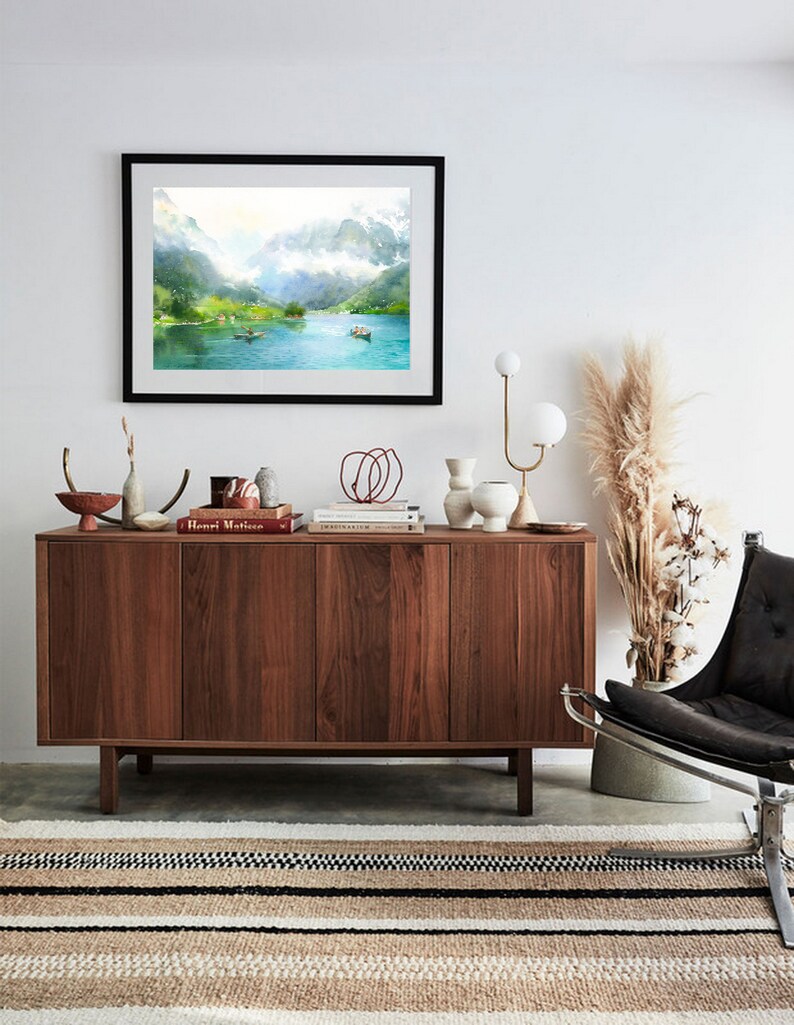 May include: A framed watercolor painting of a lake surrounded by mountains. The painting is hanging on a white wall above a wooden cabinet. The cabinet is decorated with books, vases, and other decorative items. A black leather chair with a metal frame sits in front of the cabinet.