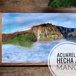 May include: A watercolor painting of a mountain range with a waterfall cascading down the side. The mountain is surrounded by clouds and a blue sky. The painting is framed in a wooden frame. The text "ACUARELA HECHA A MANO" is written in a circle below the painting.