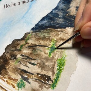 May include: A watercolor painting of a rocky cliff with green foliage. The artist is adding details to the painting with a brush. The text "Hecho a mano/Handmade" is at the top of the painting and the artist's name "Elaine Abreu" is at the bottom.