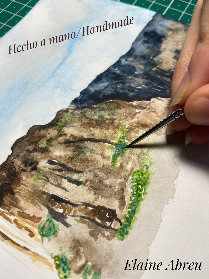 May include: A watercolor painting of a rocky cliff with green foliage. The artist is adding details to the painting with a brush. The text "Hecho a mano/Handmade" is at the top of the painting and the artist's name "Elaine Abreu" is at the bottom.