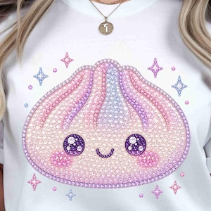 Puede incluir: Camiseta blanca con un diseño de dumpling sonriente. El dumpling es rosa, morado y azul, con detalles brillantes y acentos de estrella. El diseño tiene ojos grandes y redondos y mejillas rosadas.