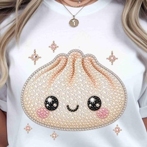 May include: White t-shirt featuring a rhinestone dumpling design with a smiling face, black eyes, and pink cheeks. The dumpling is surrounded by rhinestone star accents. The shirt is a casual, everyday wear item.