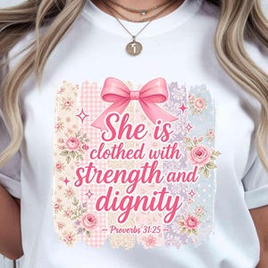 Könnte beinhalten: Weißes T-Shirt mit farbenfrohem Aufdruck. Das Design zeigt den Text "She is clothed with strength and dignity" in rosa Schrift, eine rosa Schleife, florale Elemente und den Bibelvers Sprüche 31:25.