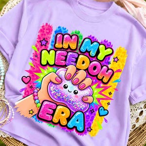 Puede incluir: Sudadera lila con un gráfico colorido que dice "IN MY NEEDOH ERA". El gráfico muestra una mano apretando una bola antiestrés con una cara sonriente. El diseño incluye estrellas, corazones y colores vibrantes.