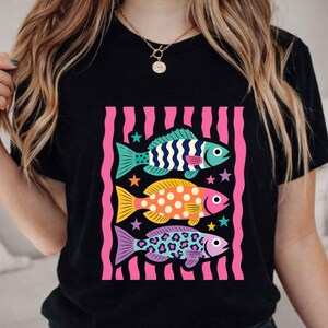 K&ouml;nnte beinhalten: Schwarzes T-Shirt mit einem farbenfrohen Grafikdesign von drei stilisierten Fischen. Die Fische sind in Blau, Orange und Lila gehalten, mit verschiedenen Mustern und umgeben von rosa Streifen und Sternen. Ein lustiges Design.