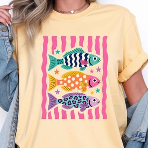 K&ouml;nnte beinhalten: Hellgelbes T-Shirt mit einer Grafik von drei stilisierten Fischen. Die Fische sind in Blau, Orange und Lila gehalten, mit Punkten und Streifen. Rosa vertikale Streifen und Sterne umgeben die Fische.