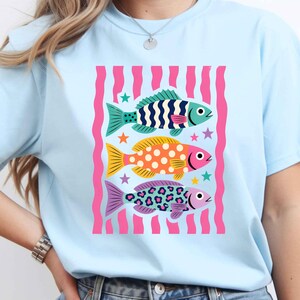 K&ouml;nnte beinhalten: Hellblaues T-Shirt mit einer Grafik von drei stilisierten Fischen. Die Fische sind in T&uuml;rkis, Orange und Lila gehalten, mit Mustern und rosa Akzenten. Das Design umfasst Sterne und einen rosa gestreiften Hintergrund. Das T-Shirt ist hellblau.