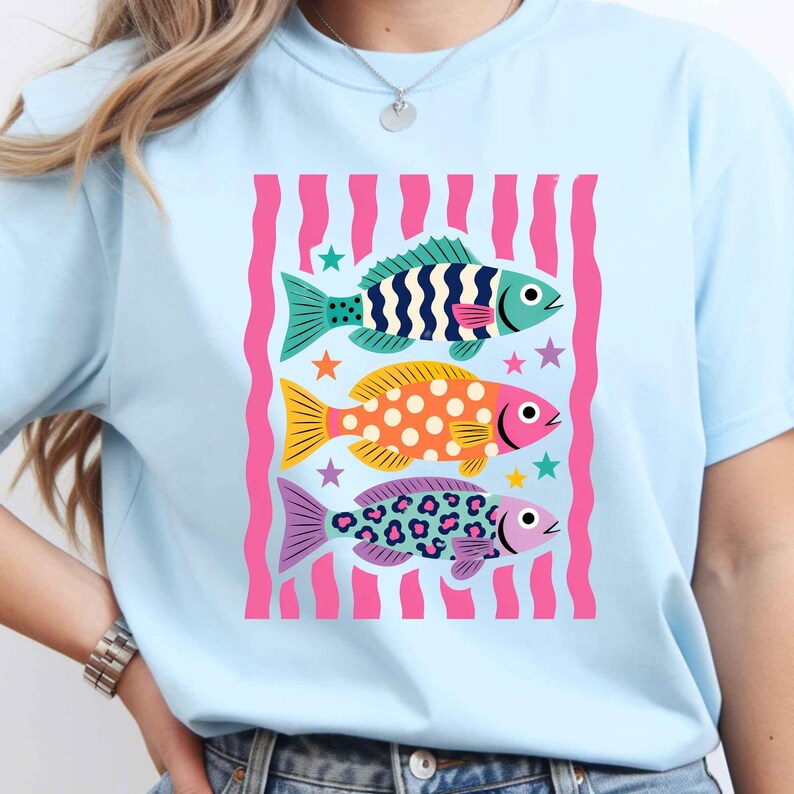 K&ouml;nnte beinhalten: Hellblaues T-Shirt mit einer Grafik von drei stilisierten Fischen. Die Fische sind in T&uuml;rkis, Orange und Lila gehalten, mit Mustern und rosa Akzenten. Das Design umfasst Sterne und einen rosa gestreiften Hintergrund. Das T-Shirt ist hellblau.