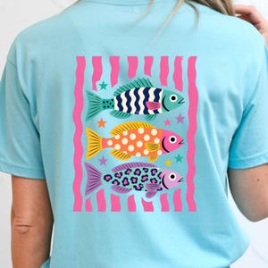 K&ouml;nnte beinhalten: Hellblaues T-Shirt mit einem farbenfrohen Fischdesign. Das Design zeigt drei Fische mit verschiedenen Mustern und Farben: blau-wei&szlig;e Streifen, orangefarbene Punkte und lila Leopardenmuster. Rosa vertikale Streifen und Sterne vervollst&auml;ndigen das Design.
