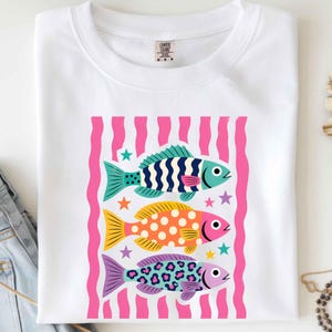 K&ouml;nnte beinhalten: Wei&szlig;es T-Shirt mit einer Grafik von drei bunten Fischen. Die Fische sind in Blau-, Gelb-, Orange- und Lilat&ouml;nen gehalten, mit einem rosa-wei&szlig; gestreiften Hintergrund. Das T-Shirt ist gefaltet.