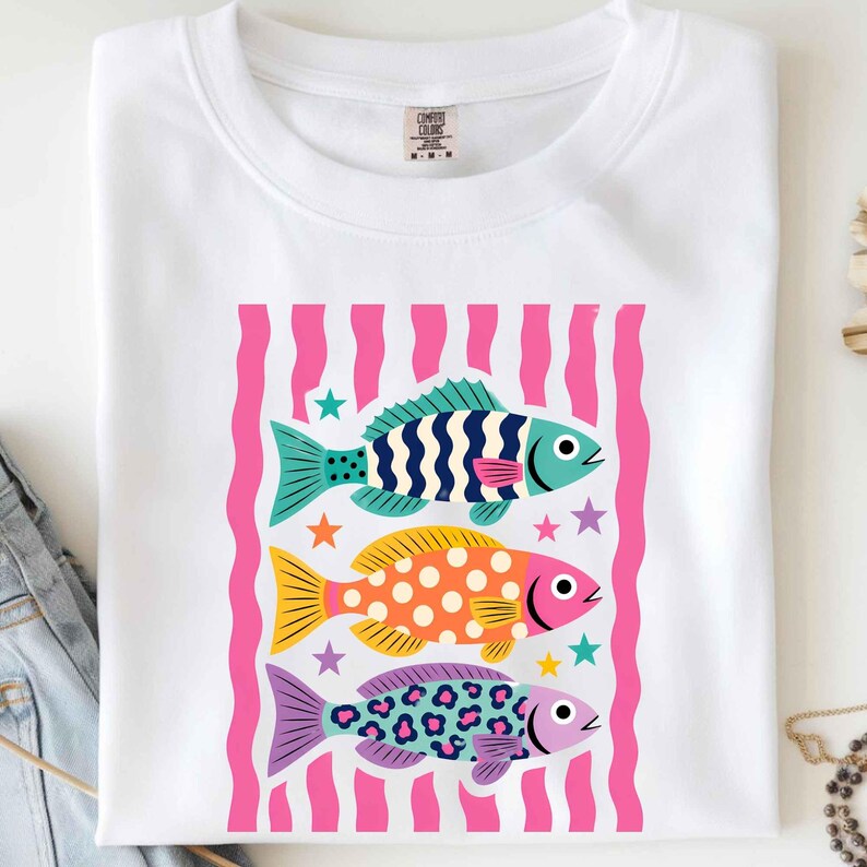 K&ouml;nnte beinhalten: Wei&szlig;es T-Shirt mit einer Grafik von drei bunten Fischen. Die Fische sind in Blau-, Gelb-, Orange- und Lilat&ouml;nen gehalten, mit einem rosa-wei&szlig; gestreiften Hintergrund. Das T-Shirt ist gefaltet.