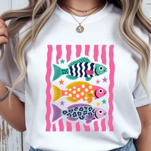 K&ouml;nnte beinhalten: Wei&szlig;es T-Shirt mit einem farbenfrohen Grafikdesign von drei stilisierten Fischen. Die Fische sind in Blau, Gelb und Lila gehalten, mit verschiedenen Mustern und umgeben von rosa Streifen und Sternen. Ein l&auml;ssiges, lustiges Design.