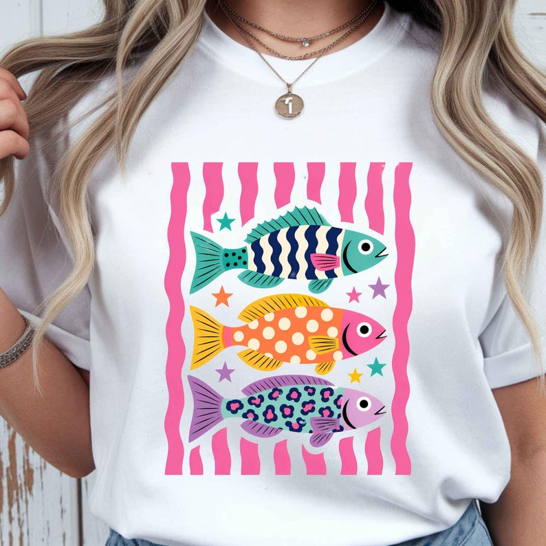 K&ouml;nnte beinhalten: Wei&szlig;es T-Shirt mit einem farbenfrohen Grafikdesign von drei stilisierten Fischen. Die Fische sind in Blau, Gelb und Lila gehalten, mit verschiedenen Mustern und umgeben von rosa Streifen und Sternen. Ein l&auml;ssiges, lustiges Design.