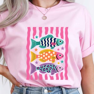 K&ouml;nnte beinhalten: Hellrosa T-Shirt mit einer Grafik von drei bunten Fischen. Die Fische sind in Blau, Gelb und Lila gehalten und weisen verschiedene Muster auf. Das Design befindet sich vor einem rosa-wei&szlig; gestreiften Hintergrund.