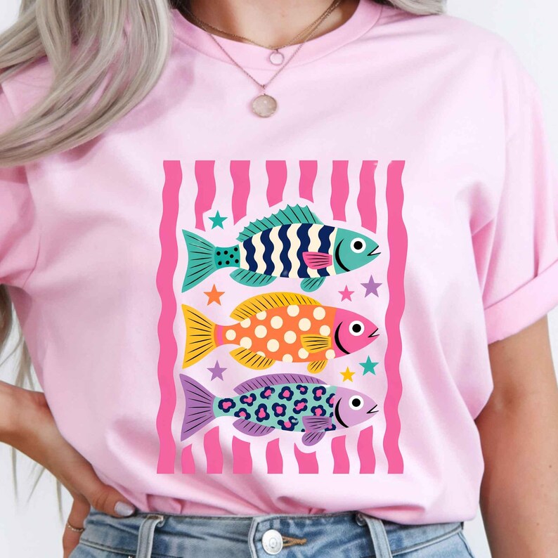 K&ouml;nnte beinhalten: Hellrosa T-Shirt mit einer Grafik von drei bunten Fischen. Die Fische sind in Blau, Gelb und Lila gehalten und weisen verschiedene Muster auf. Das Design befindet sich vor einem rosa-wei&szlig; gestreiften Hintergrund.
