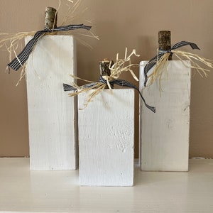 May include: Three white wooden pumpkins with a rustic, farmhouse style. Each pumpkin is decorated with a natural twig stem, burlap, and black and white checkered ribbon.